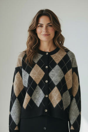 A woman stands indoors, smiling. She wears a white button-down shirt, a black buttoned skirt, and the HARTFORD - MEDLIAA ARGYLE WOOL CARDIGAN by HARTFORD in black, brown, and gray tones. The background is plain and softly lit.