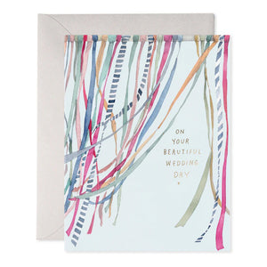 The E. FRANCES PAPER - WEDDING RIBBONS GREETING CARD features a lively watercolor painting with vibrant blue, pink, and green vertical ribbons, complemented by gold text that reads "On your beautiful wedding day." It comes with a beige envelope.