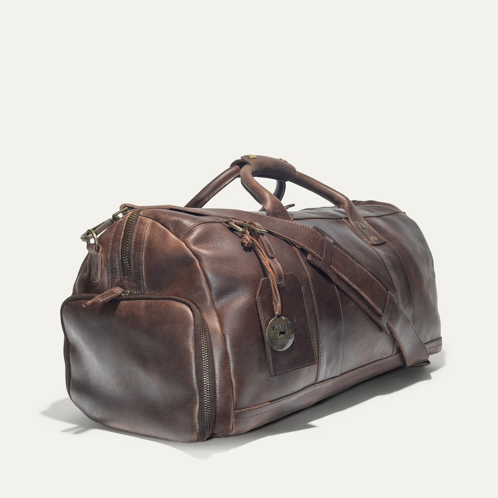 A brown leather duffel bag with handles and a shoulder strap, featuring a front pocket, side zippers, and a small round tag, set against a plain white background.