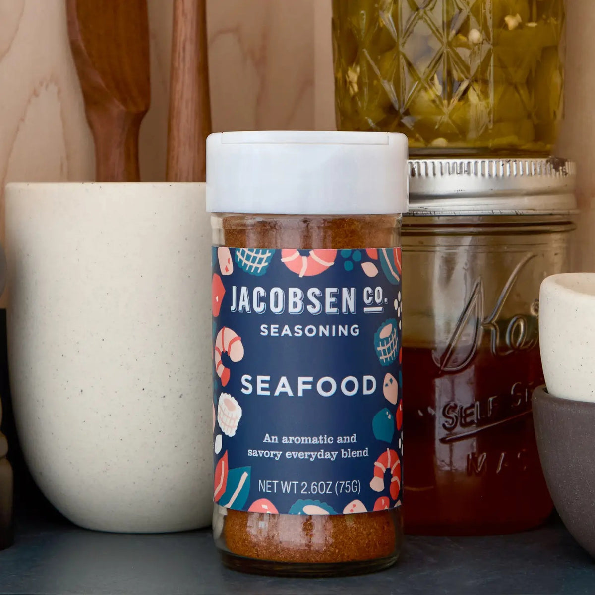 A clear spice jar with a white lid from JACOBSEN SALT CO., featuring a vibrant seafood-themed label for the JACOBSEN SALT - SEAFOOD SEASONING. The jar contains an orange-red blend made with sea salt, herbs, and spices—perfect for elevating your favorite seafood dishes.