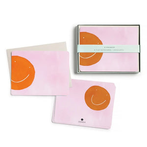 E. FRANCES PAPER’s "Flat Notes Orange Smiley" set features a simple orange smiley face on pink, paired with a plain white envelope—ideal for sending illustrated notecards or thoughtful, handwritten notes.