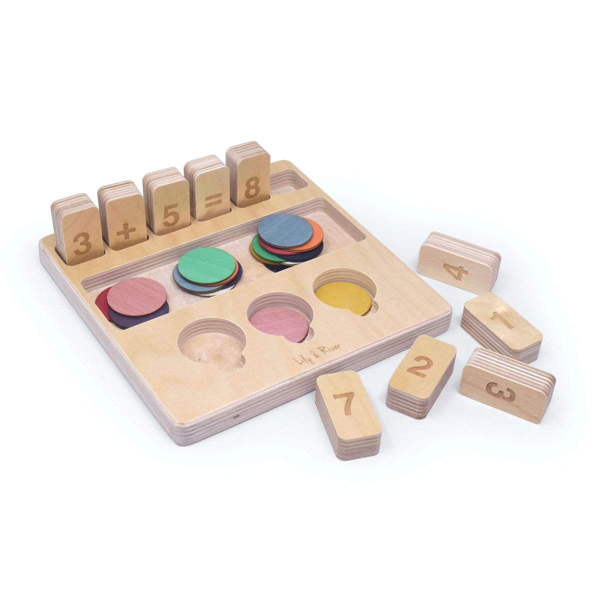The "LITTLE NUMBERS GAME SET" by LILY &amp; RIVER is a wooden educational toy influenced by Montessori principles. It features number blocks and colored disks on a white surface. The blocks display a simple math equation (2 + 2 = 4), functioning like a children's calculator. In the background, there is a light-colored fabric bag with the text "Lily &amp; River" partially visible.
