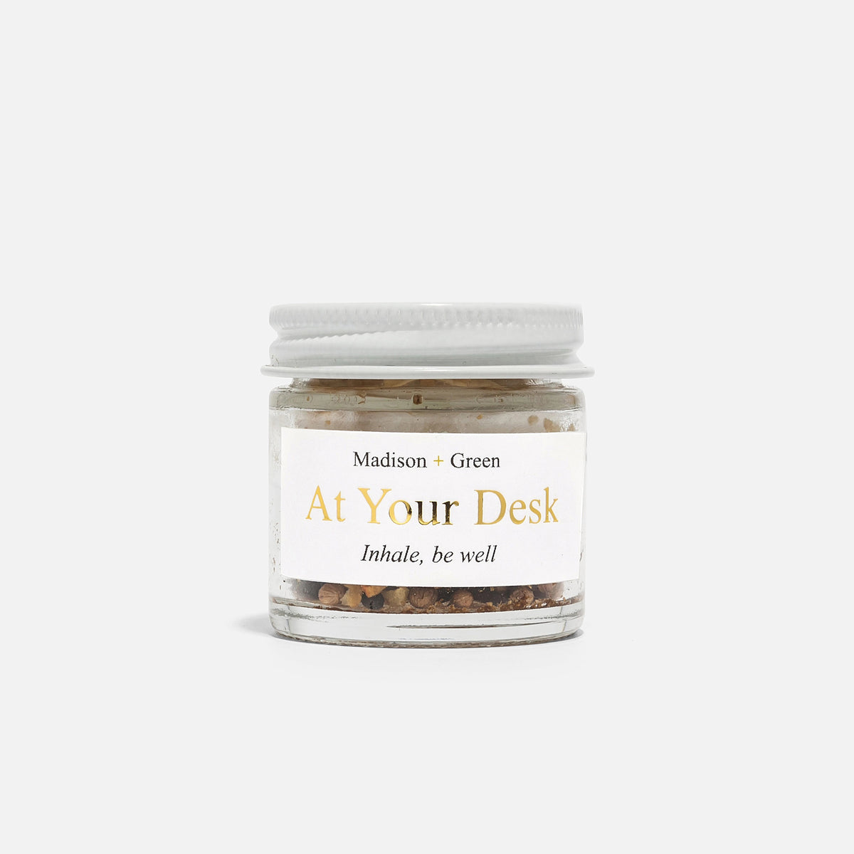 A small glass jar with a white lid labeled "MADISON AND GREEN - AT YOUR DESK FOCUS BOOST AROMATHERAPY INHALER" by MADISON GREEN contains a blend of dried herbs for stress relief, shown against a plain white background.