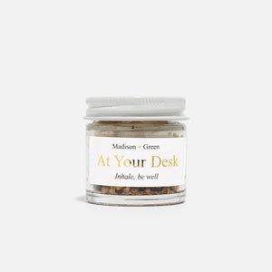 A small glass jar with a white lid labeled "MADISON AND GREEN - AT YOUR DESK FOCUS BOOST AROMATHERAPY INHALER" by MADISON GREEN contains a blend of dried herbs for stress relief, shown against a plain white background.
