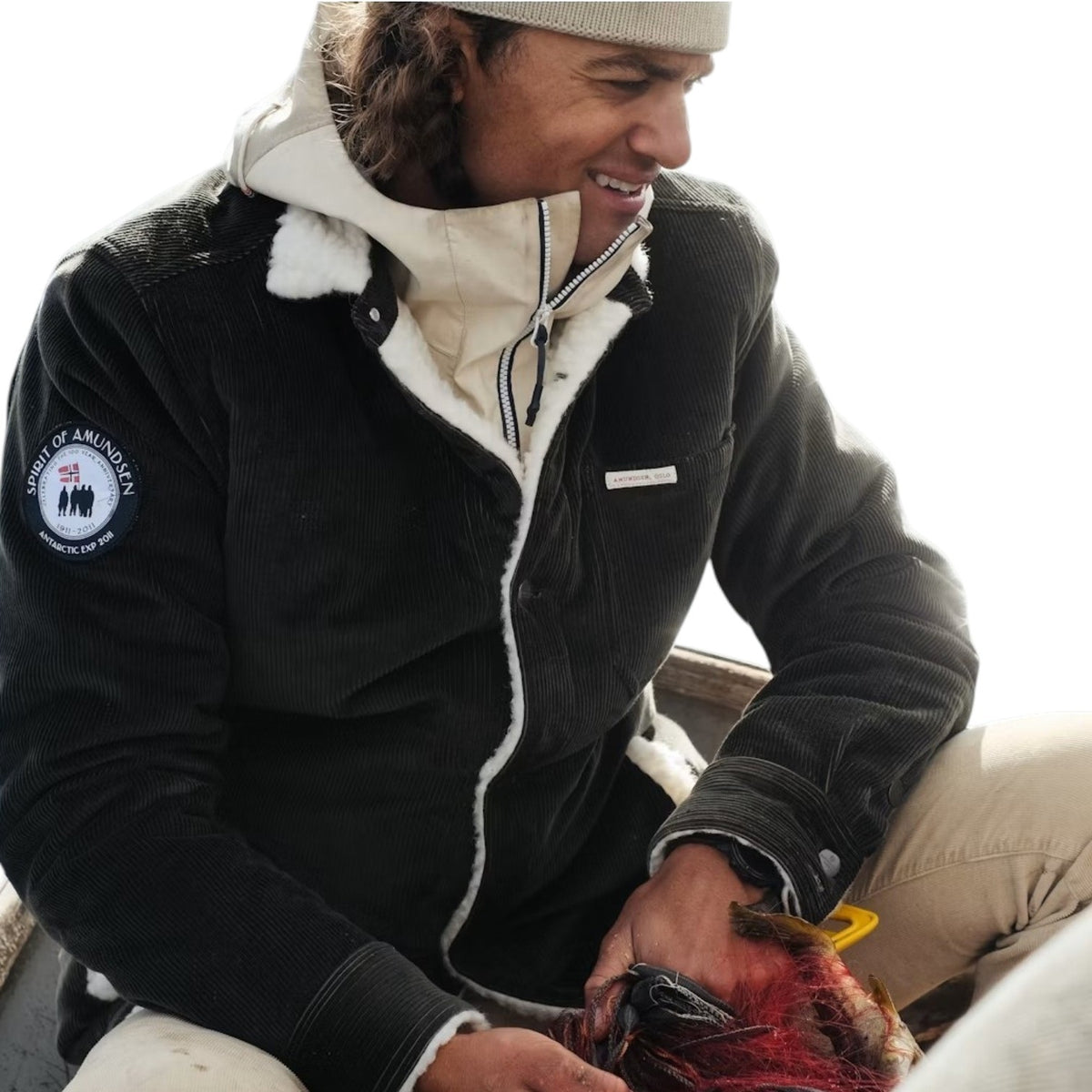A person in a warm jacket and beanie smiles while holding a red-feathered bird, possibly on a boat, wearing the AMUNDSEN SPORTS - HARVESTER OVERSHIRT.