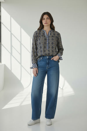 A person with long brown hair, seen from behind, wears the STORIATIPIC MARGOT BLOUSE IN BLUE by STORIATIPIC—a patterned cotton shirt featuring a mix of vertical stripes and diamond shapes in blue and beige tones—paired with blue jeans. The blouse is suitable for a gentle machine wash at 30 degrees.