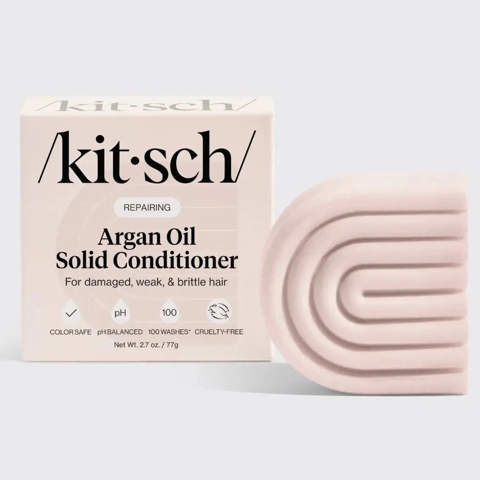 A pale pink, geometric KITSCH Argan Oil Conditioner Bar is pictured beside its box, formulated for damaged, weak, brittle hair. It’s cruelty-free, reduces frizz with argan oil, and lasts up to 100 washes.