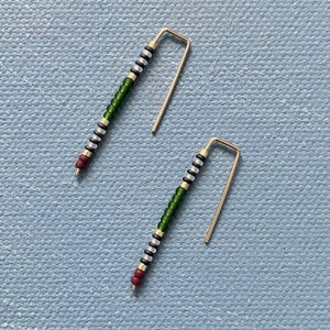 The ALICE RISE - BIG QUILL EARRINGS IN FOREST GREEN by ALICE RISE feature 14k gold-filled hooks and small red, green, black, and white beads. Displayed parallel on a textured light blue background, their hooks face opposite directions.