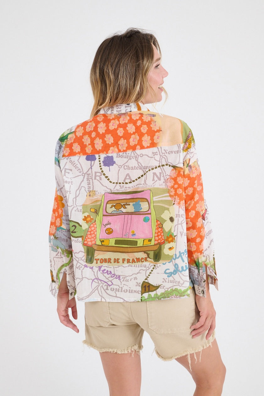 A smiling woman with long wavy hair wears the STORIATIPIC - SUZIE GABIE BLOUSE, a colorful off-the-shoulder cotton top with map and floral patterns, paired with beige frayed shorts against a plain white background.