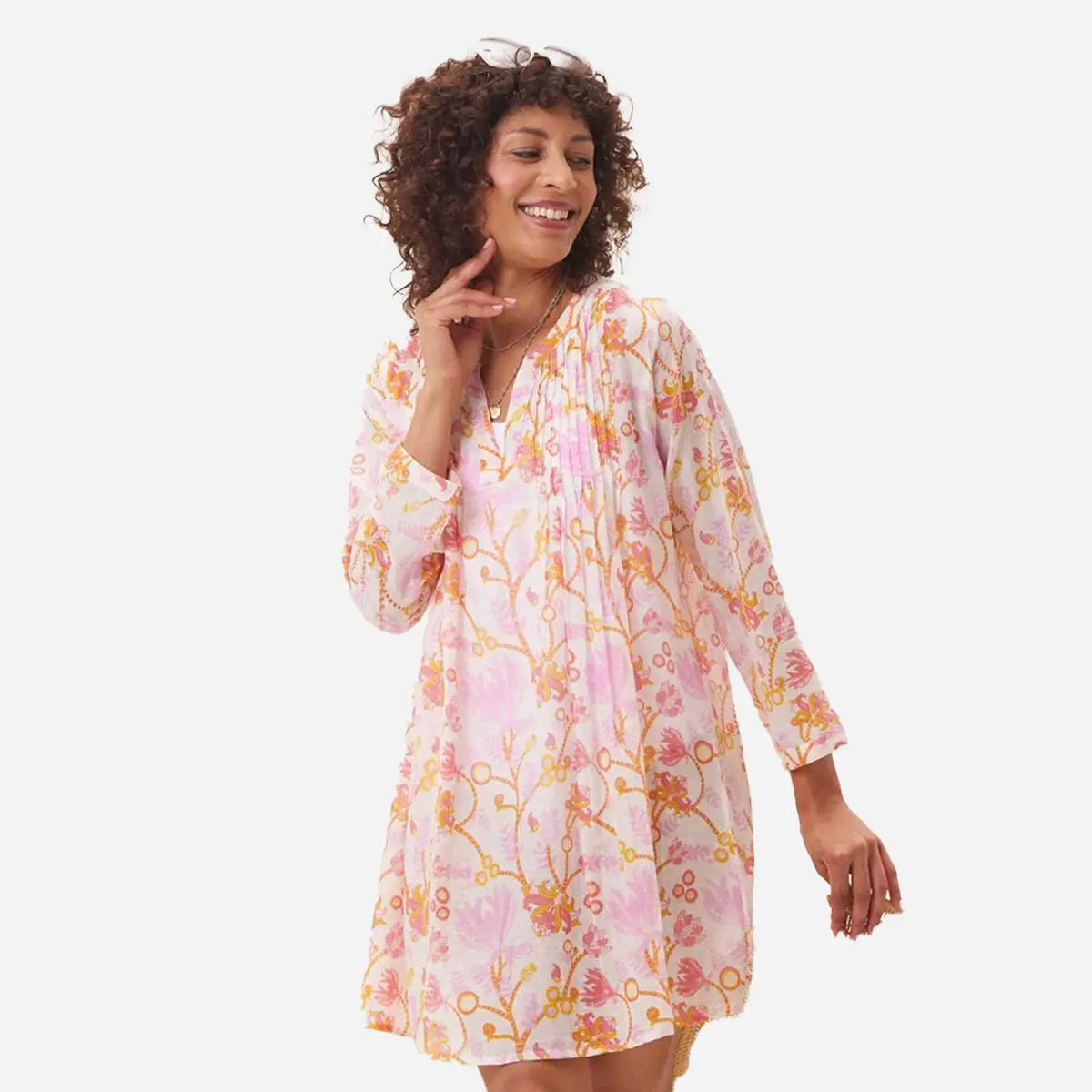 A person wears the ROCKFLOWERPAPER GEMMA Pink Pintuck Beach Cover Up—a lightweight long-sleeved blouse with a pink and yellow floral pattern and split neckline—styled with layered delicate necklaces.