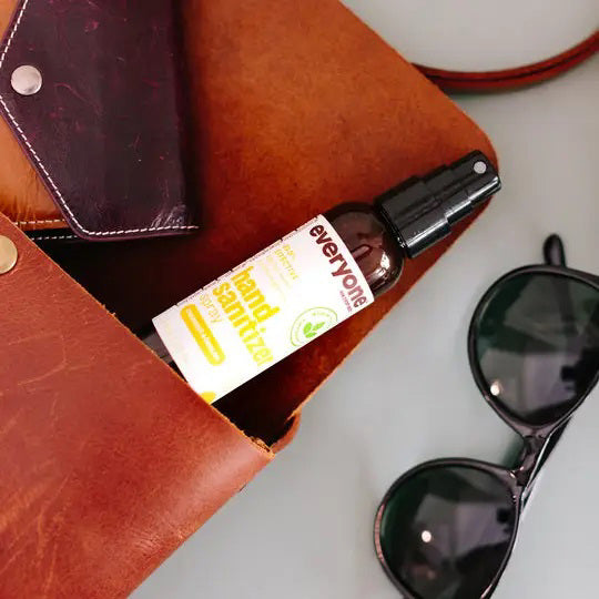 A brown leather bag is open, showing a bottle of Everyone For Everybody Coconut &amp; Lemon Hand Sanitizer Spray with 62% alcohol and a wallet inside. Adjacent to the bag are black sunglasses. The scene suggests preparedness for hygiene and travel or outdoor activities.