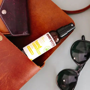 A brown leather bag is open, showing a bottle of Everyone For Everybody Coconut & Lemon Hand Sanitizer Spray with 62% alcohol and a wallet inside. Adjacent to the bag are black sunglasses. The scene suggests preparedness for hygiene and travel or outdoor activities.
