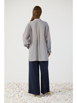 A woman wears the ELEH - BLOUSE WITH DRAPE FRONT in light gray, featuring relaxed tailoring, long sleeves, a collar, front tie detail, and an asymmetrical hem. It pairs effortlessly with dark pants for a chic look.