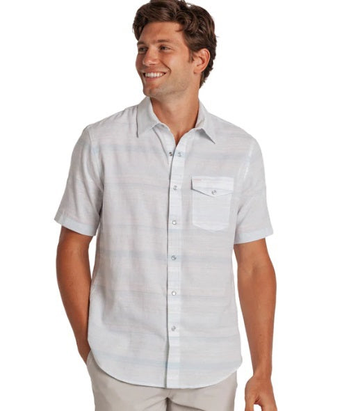A man stands outdoors in sunlight wearing the CRIQUET HEMP STRIPE PEARL SNAP SHIRT IN LIGHT BLUE, featuring subtle horizontal stripes, short sleeves, a front pocket, and partial view of his bearded face. Brand: CRIQUET.