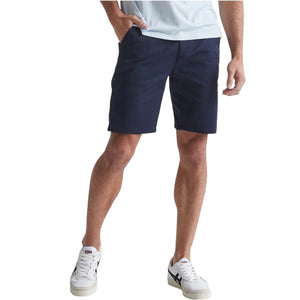 A man in a light blue T-shirt, DUER Live Free Journey Shorts in Sapphire, and white sneakers with black details stands with one hand in his pocket against a plain light background.