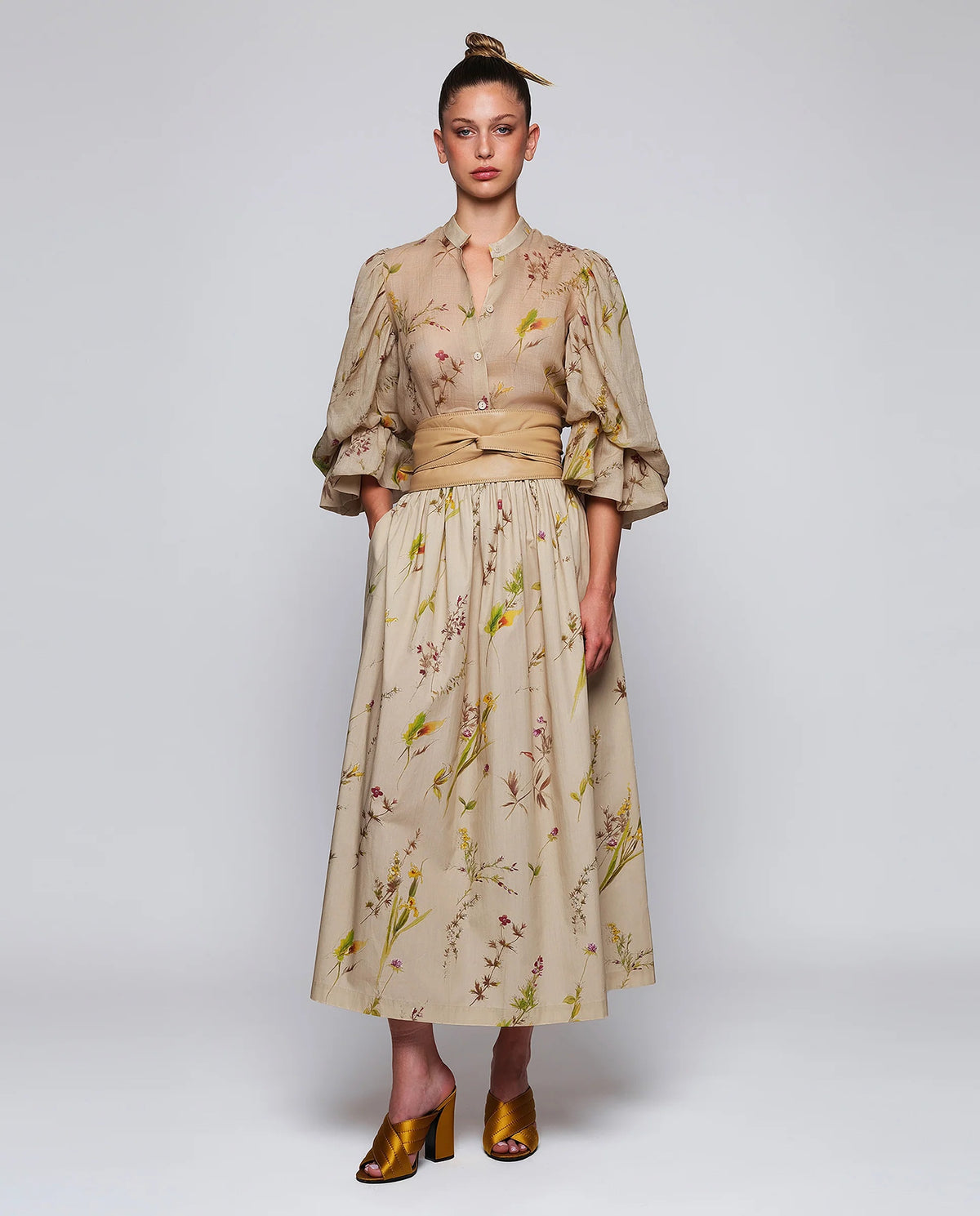 A woman stands against a plain background wearing the MIRTO 1956 Beige Floral Print Midi Skirt, paired with puffed three-quarter sleeves, a wide tan belt, and brown open-toe heels. Her hair is styled in a high bun with a decorative accessory.