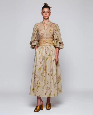 A woman stands against a plain background wearing the MIRTO 1956 Beige Floral Print Midi Skirt, paired with puffed three-quarter sleeves, a wide tan belt, and brown open-toe heels. Her hair is styled in a high bun with a decorative accessory.