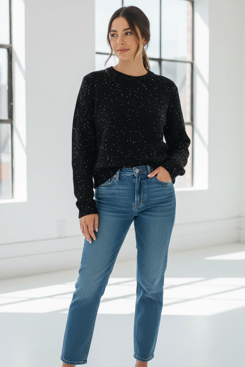 A woman with long dark hair, dressed in the HINSON WU - QUEENIE LONG SLEEVE SEQUIN SWEATER IN BLACK by HINSON WU and blue jeans, stands against a plain light background, smiling gently at the camera.