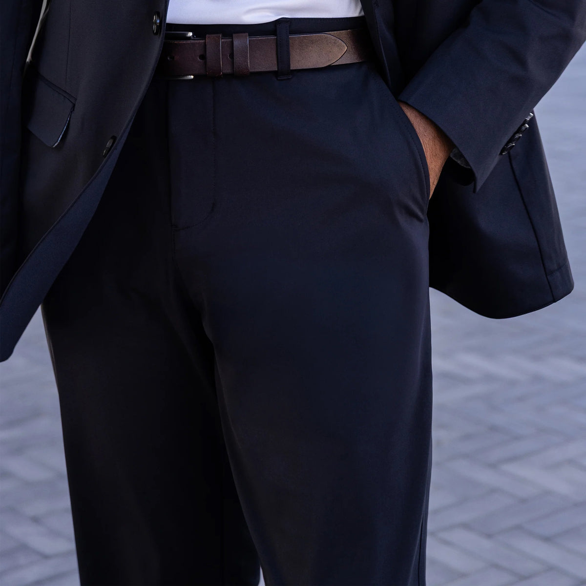 Close-up of a person wearing COLLARS AND CO’s Maverick Performance Pants in Black, paired with a dark suit jacket, white shirt, and brown belt. The open jacket reveals the inner lining and label. Light brick background.