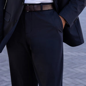 Close-up of a person wearing COLLARS AND CO’s Maverick Performance Pants in Black, paired with a dark suit jacket, white shirt, and brown belt. The open jacket reveals the inner lining and label. Light brick background.