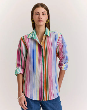 A person confidently poses with hands on hips against a light background, wearing the HINSON WU - HALSEY LONG SLEEVE LINEN SHIRT IN MULTI STRIPE and blue jeans.