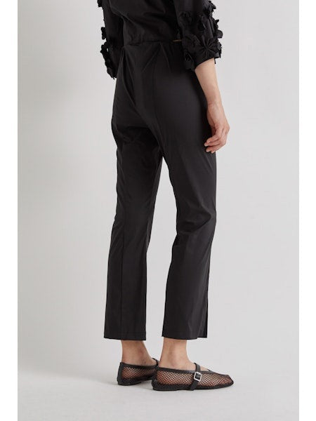 A person stands against a plain background wearing MEIMEIJ FROTN HEM SPLIT COTTON PANT—black, cropped, ankle-length pants with small slits. They also wear a black textured-sleeve top and black shoes. Their face is not visible in the image.
