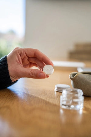A hand pours liquid from a small ceramic pitcher into a glass jar filled with a white, fluffy substance atop a wooden surface, demonstrating the preparation of essential oil tablets for use with the MADISON AND GREEN - REST YOUR EYES AT THE DESK aromatherapy eye covers or reusable eye mask by MADISON GREEN.