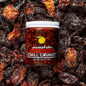A jar of MOMOFUKU - EXTRA SPICY CHILI CRUNCH is shown against a white background. The jar contains 150 grams (5.3 ounces) of smoky spiciness, with a vibrant red color and visible Mexican chilis, highlighted by a yellow peach graphic above the brand name, MOMOFUKU.