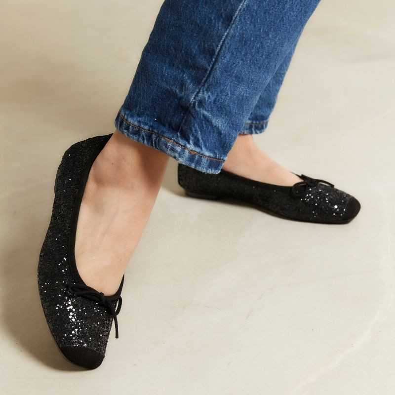 The REQINS Harmony Sequin Ballerina Flats in Noir feature black sequins, black toe caps, and small bows—bringing a touch of everyday luxury against a plain light background.