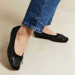 The REQINS Harmony Sequin Ballerina Flats in Noir feature black sequins, black toe caps, and small bows—bringing a touch of everyday luxury against a plain light background.