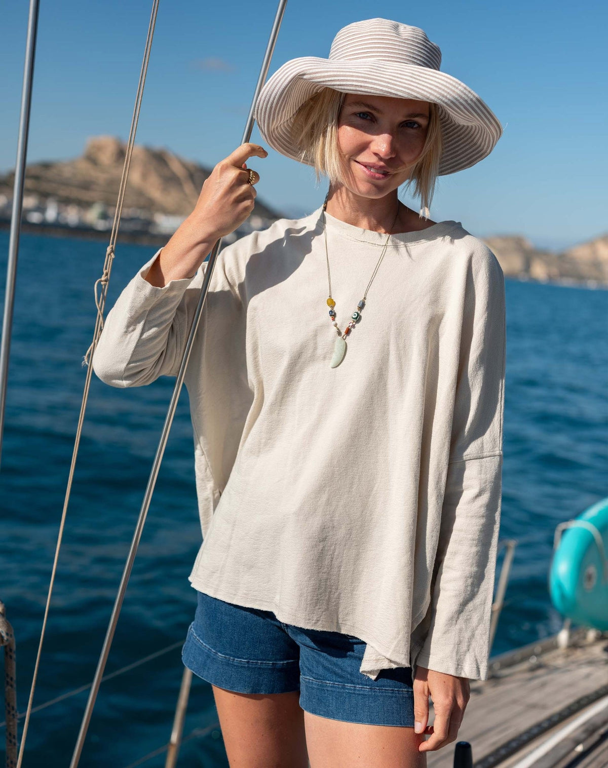 A woman stands barefoot in sunlight, smiling with one hand in her pocket while wearing the MERSEA - CATALINA RUSTIC TEE IN BONE paired with blue denim shorts, in a minimalist room.