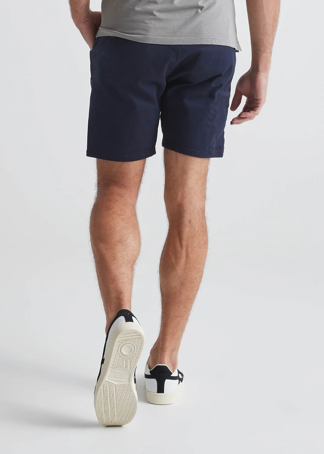 A man in a light blue T-shirt, DUER Live Free Journey Shorts in Sapphire, and white sneakers with black details stands with one hand in his pocket against a plain light background.