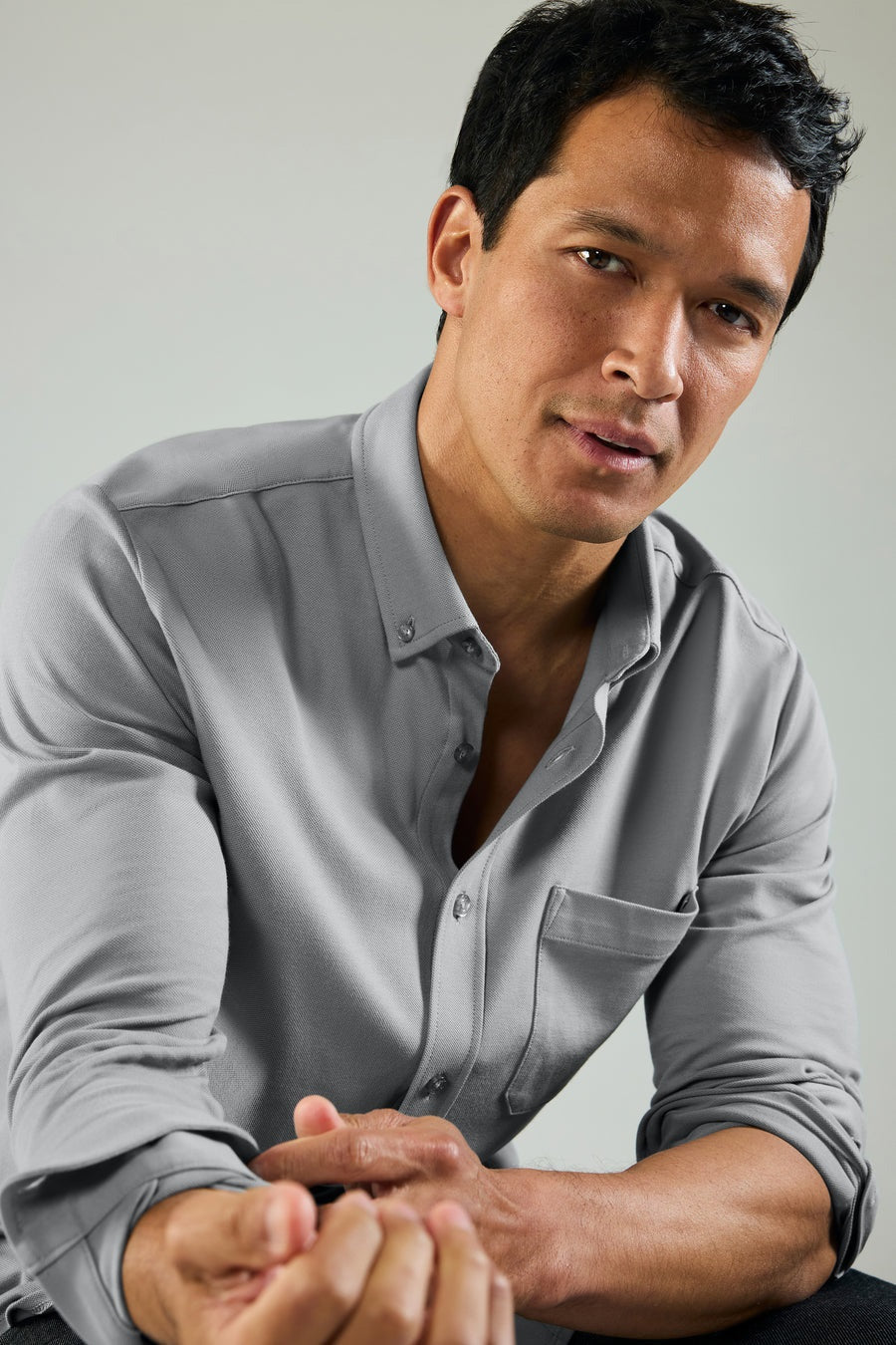 A man with short dark hair wears the MACK WELDON Silver Pique Long Sleeve Button Up Shirt in True Navy, standing against a plain light background with a neutral expression and his hands together in front of him.