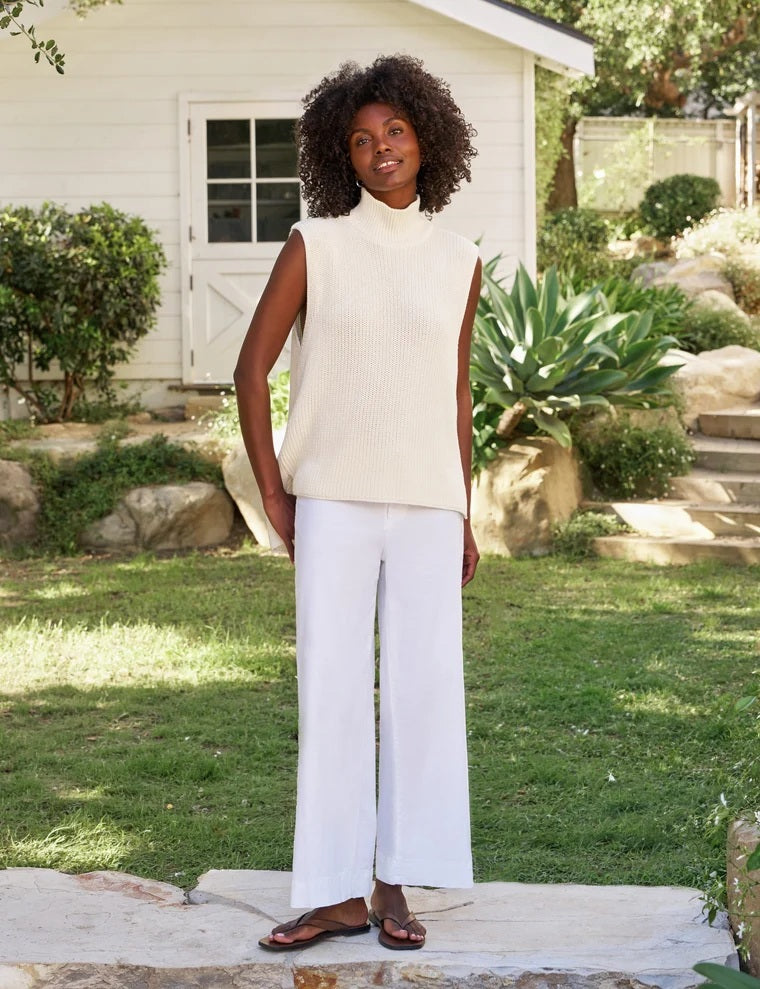 Wearing the FRANK & EILEEN MONTECITO Shell Mock Neck Sweater Shell in Ivory, a person with curly hair and white pants stands outdoors with their back to the camera, in front of a white building surrounded by greenery.