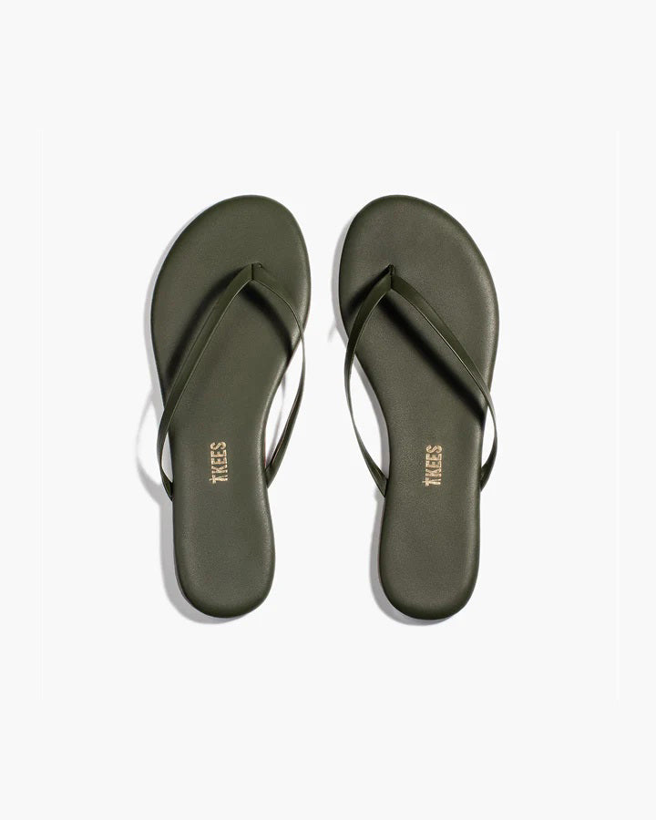 The TKEES INC LILY LINER SLIPPER FOREST is a dark green flip-flop sandal with thin straps and a cushioned insole for comfort. The brand name &quot;TKEES&quot; is printed in gold near the heel on the sole. Photographed from above on white background.