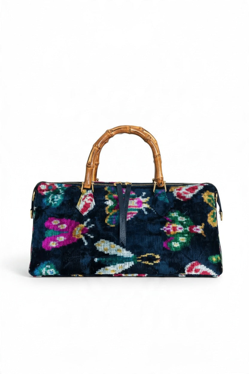 The BALDIZ GOLDIE BAMBOO BAG IN OXFORD BLUE BUG is a handcrafted rectangular bag in dark blue fabric featuring a colorful moth pattern and bamboo handles.