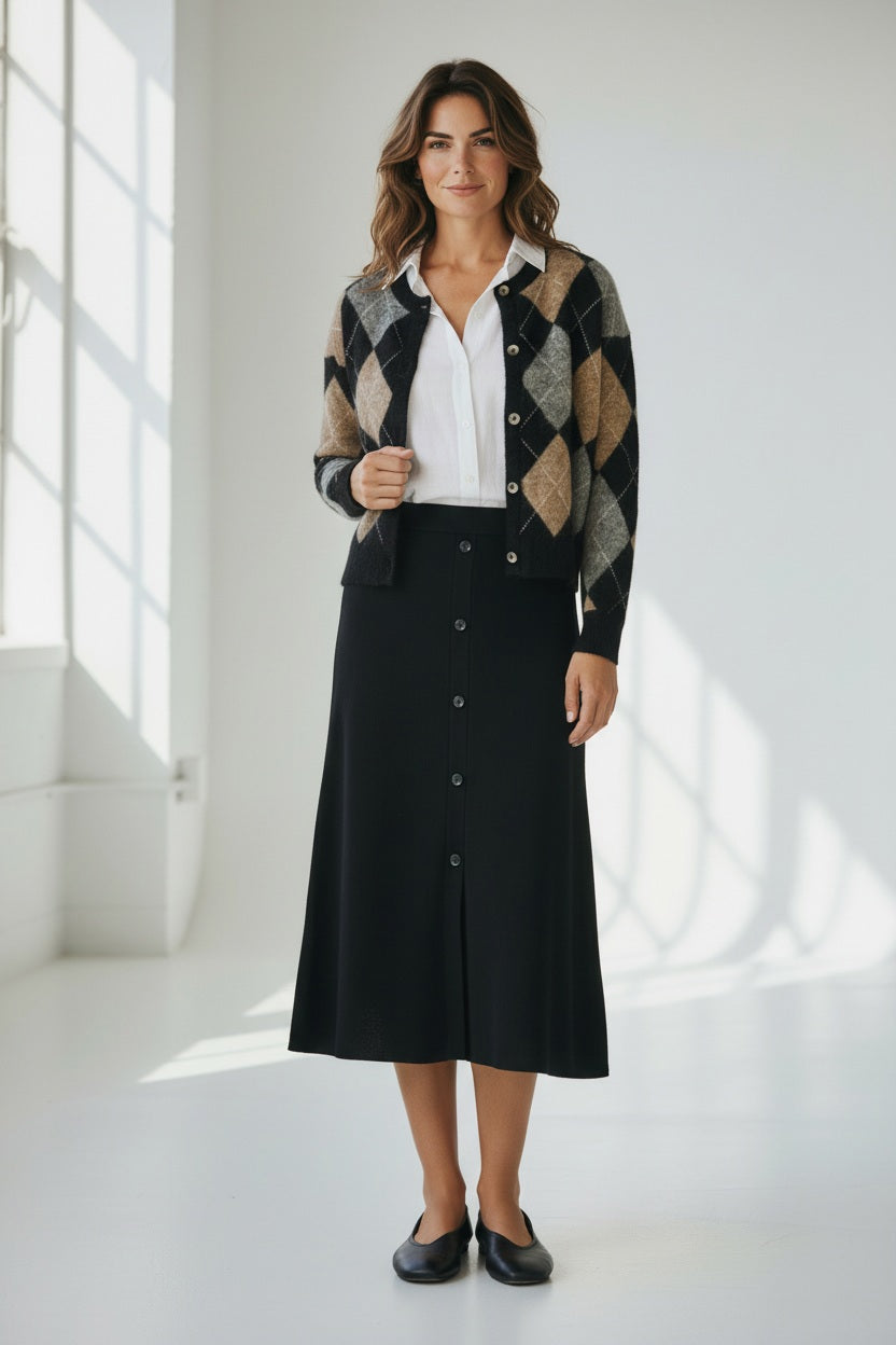 A woman stands indoors, smiling. She wears a white button-down shirt, a black buttoned skirt, and the HARTFORD - MEDLIAA ARGYLE WOOL CARDIGAN by HARTFORD in black, brown, and gray tones. The background is plain and softly lit.