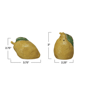 A yellow, lemon-shaped bowl of grains sits on a light surface. In front are CREATIVE COOP STONEWARE LEMON-SHAPED SALT AND PEPPER SHAKERS with green leaves, adding charm to your kitchen décor as grains scatter nearby.