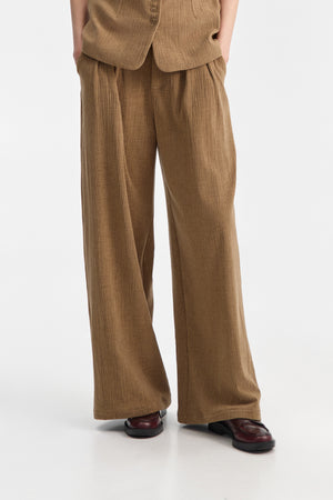 A person wearing ELEH CLASSIC WIDE LEG TROUSERS and a matching brown corduroy jacket, paired with brown shoes and large sunglasses, stands against a plain white background with hands in pockets.