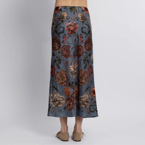 A person wearing the SABINA SAVAGE England's "Ring a Ring O' Roses" silk bias skirt, featuring a botanical print with red, orange, and cream flowers on a blue background, stands against a plain white wall. The elegant skirt is paired with beige flat shoes.