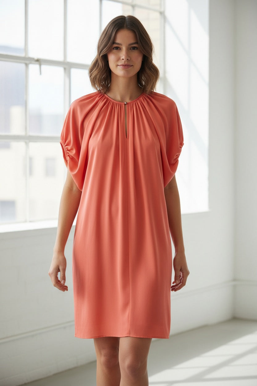A woman stands indoors by a large window, dressed in the MOMONI - OLIVIA DRESS from MOMONI. The elegant white dress features gathered sleeves reminiscent of silk satin crepe. She pairs it with brown ankle boots, while the minimal background includes a gray wall and a partially visible plant.