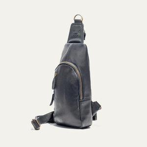The WILL LEATHER GOODS - ZIP AROUND SLING BAG IN BLACK is a full-grain leather sling bag with gold zippers and buckle, small front pocket, adjustable strap, and top hanging loop, shown against a plain white background.
