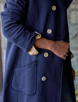 A woman stands indoors by a window, smiling with her hands in the pockets of a FRANK & EILEEN Belfast Sweatshirt Trench Coat in Classic Navy from the California Heritage Collection. She wears a tan shirt, wide-leg blue jeans, and brown boots. A wooden stool is positioned behind her.
