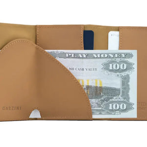 The GARZINI - SLIM TRIFOLD RFID-blocking leather wallet in camel brown is shown open, displaying a blue card, a white card, and a large “NO CASH VALUE” play bill. “GARZINI” is embossed on the left side. Includes metal card holder and coin pocket.