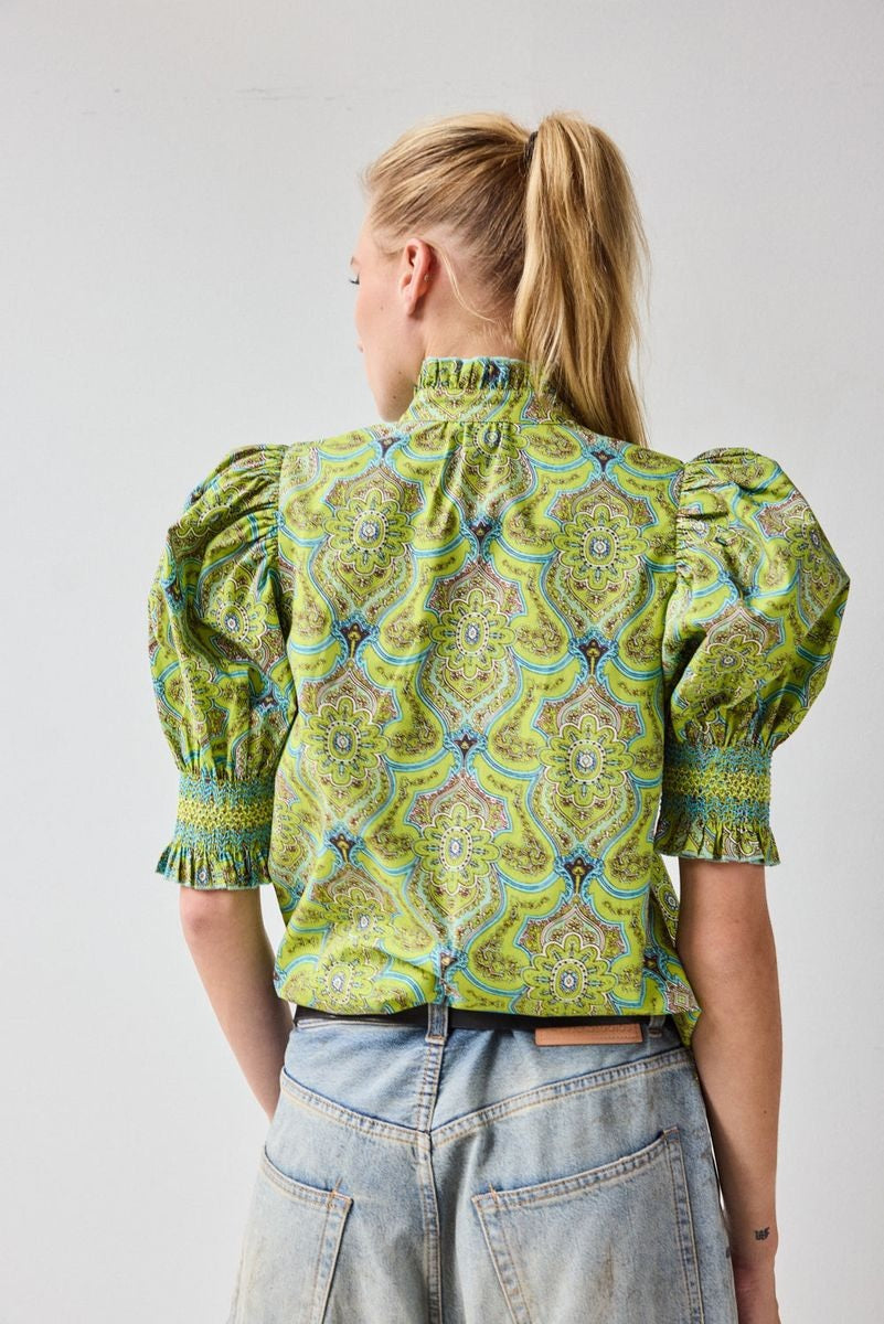 A woman with blonde hair in a ponytail wears the HOPE FOR FLOWERS Short Sleeve Smocked Shirt in Green Foulard, made of organic cotton, paired with faded blue jeans, standing with her back to the camera against a plain light background.