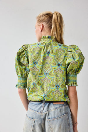 A woman with blonde hair in a ponytail wears the HOPE FOR FLOWERS Short Sleeve Smocked Shirt in Green Foulard, made of organic cotton, paired with faded blue jeans, standing with her back to the camera against a plain light background.