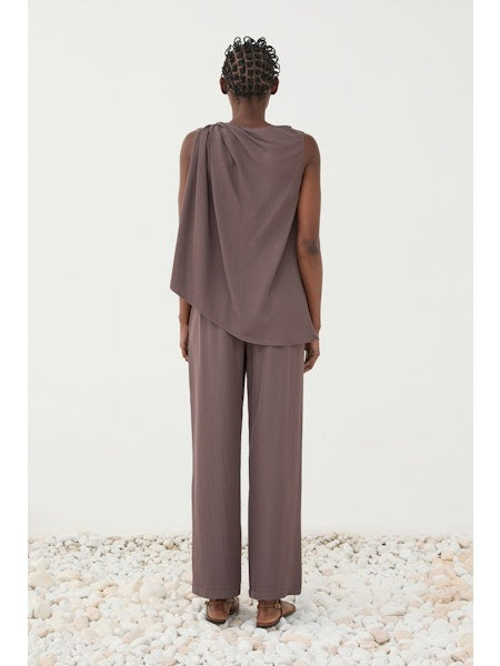 A person wears the ELEH TOP WITH SIDE KNOT, a sleeveless mauve, loose-fit blouse with an asymmetrical neckline and knot detail, paired with matching wide-leg pants. One hand is in their pocket against a plain light background.