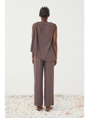 A person wears the ELEH TOP WITH SIDE KNOT, a sleeveless mauve, loose-fit blouse with an asymmetrical neckline and knot detail, paired with matching wide-leg pants. One hand is in their pocket against a plain light background.