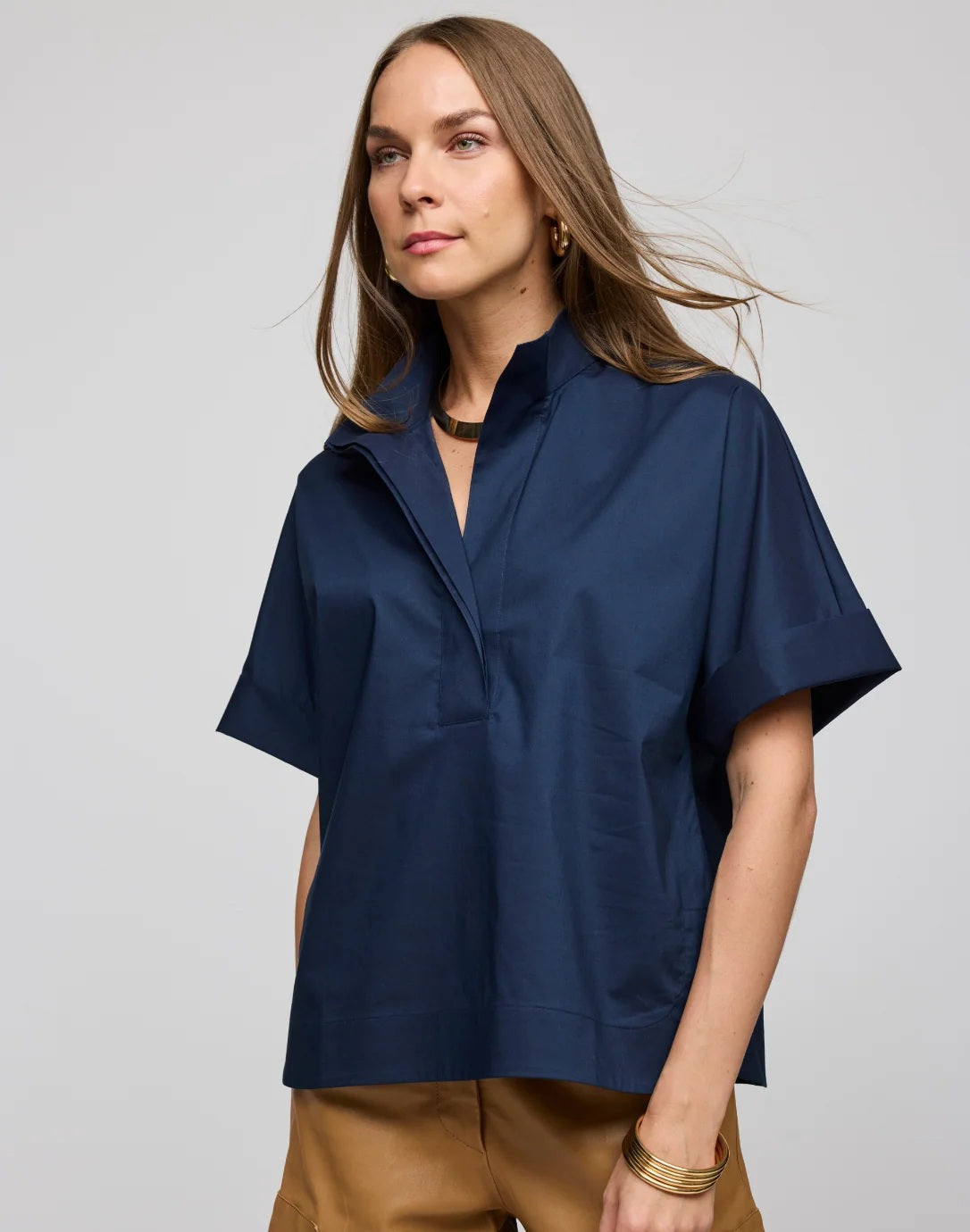 A woman with long brown hair is seen wearing the HINSON WU Cara Elbow Sleeve Shirt in Stretch Polished Cotton Navy—a loose-fitting, oversized short-sleeve shirt—paired with tan pants and gold jewelry, standing against a plain light gray background.