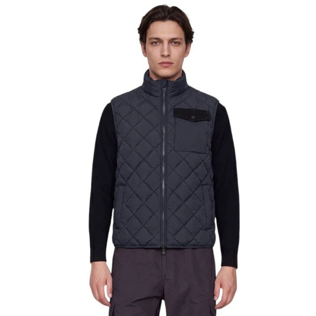 A person stands facing forward, wearing the SAVE THE DUCK - FABRICE Puffer Vest in Blue Black from SAVE THE DUCK, featuring recycled Plumtech insulation, layered over a black long-sleeve shirt and gray pants. They have a neutral facial expression and are posed against a plain white background.